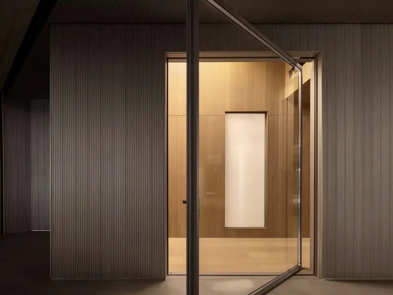 Dark, moodily lit interior corridor shot of a full-height glass pivot door swung open on its central vertical pivot, revealing a warm timber-panelled room beyond.