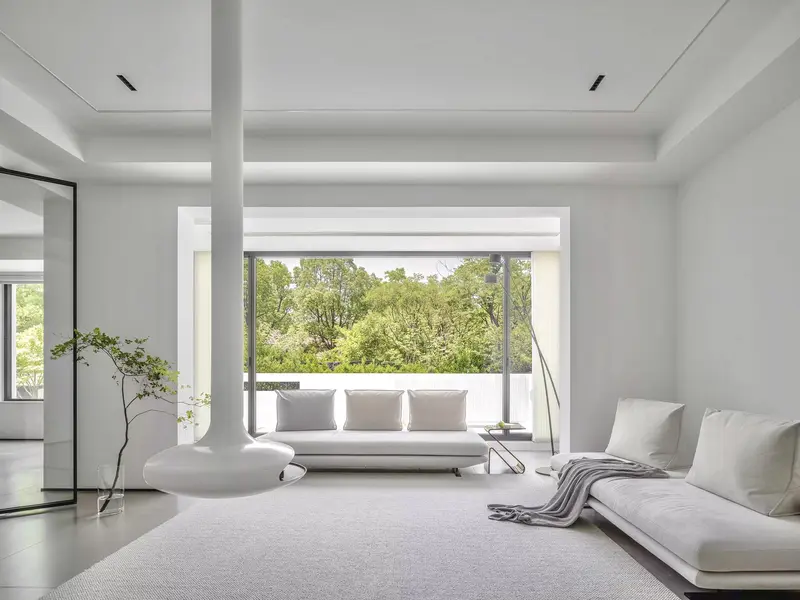 All-white minimalist living room with a floor-lamp and sectional sofa beside a large glass window/door opening onto a tree-filled terrace.