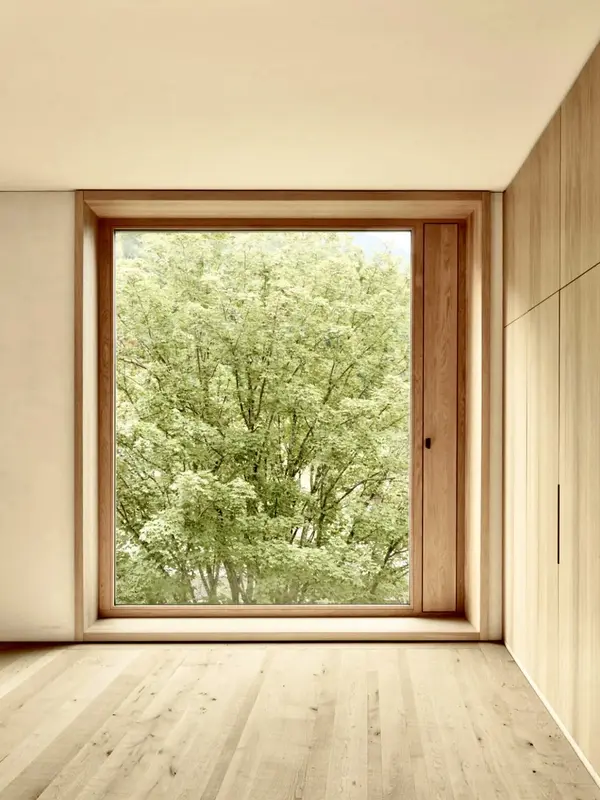 Wood-framed picture window with a narrower hinged ventilation flap beside it, viewed from an empty room with light timber flooring; large tree visible outside.